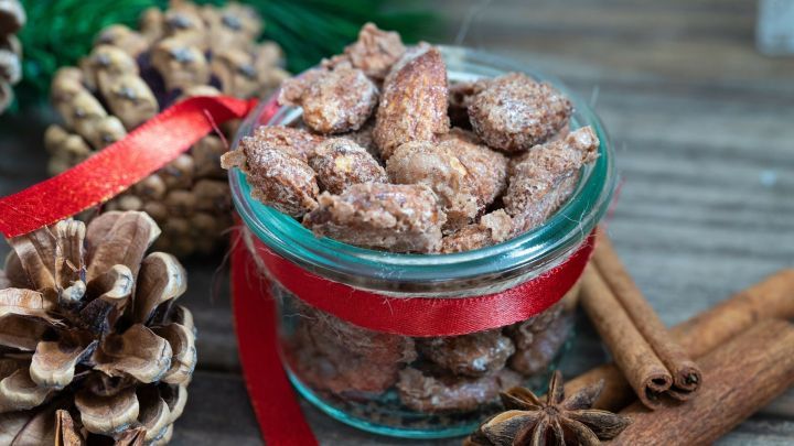 In einem klaren Glasgef&auml;&szlig; sind gebrannte Mandeln sichtbar, umgeben von Zimtstangen und Tannenzapfen.