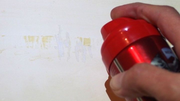 Ein Hand h&auml;lt einen roten Deo-Roller und entfernt Klebereste vom doppelseitigen Klebeband auf einer wei&szlig;en Oberfl&auml;che.