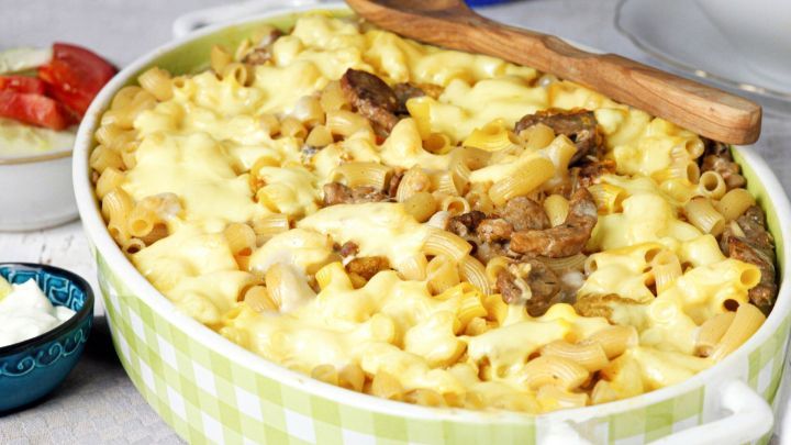 Ein Auflauf mit Gyros, Makkaroni und geschmolzenem K&auml;se wird in einer gr&uuml;nen Auflaufform serviert, umgeben von Beilagen.