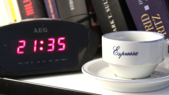 Ein Wecker zeigt 21:35 Uhr, w&auml;hrend eine Espressotasse auf einem Tisch neben B&uuml;cherregalen steht, was Einschlafst&ouml;rungen symbolisiert.