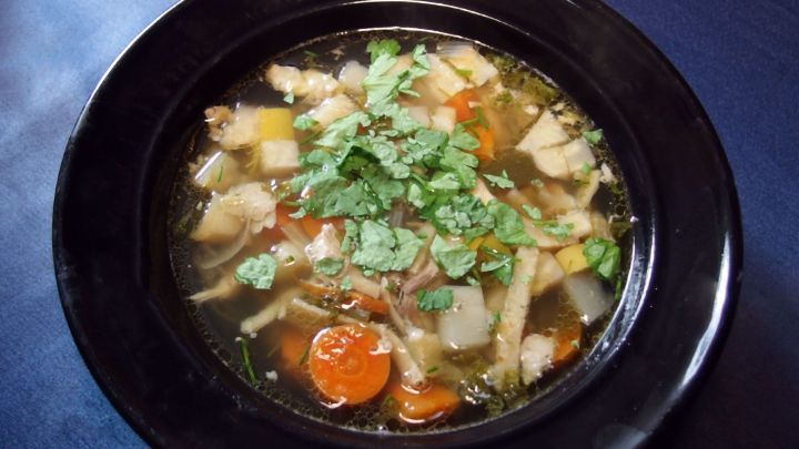 Ein schwarzer Teller mit H&uuml;hnersuppe, gef&uuml;llt mit Gem&uuml;se und frischem Koriander, auf einem blauen Tisch.