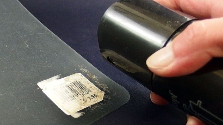 Eine Hand h&auml;lt eine Haarspray-Dose, w&auml;hrend sie Stickerreste von einer glatten Fl&auml;che entfernt.