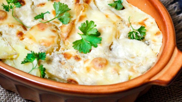 Ein goldbrauner Kartoffelauflauf mit Milch, garniert mit frischem Koriander, steht in einer Keramikform auf einem rustikalen Untergrund.