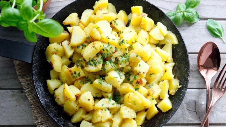 In einer schwarzen Pfanne liegen goldbraun angebratene Kartoffelst&uuml;cke, umgeben von frischem Basilikum auf einem Holztisch.