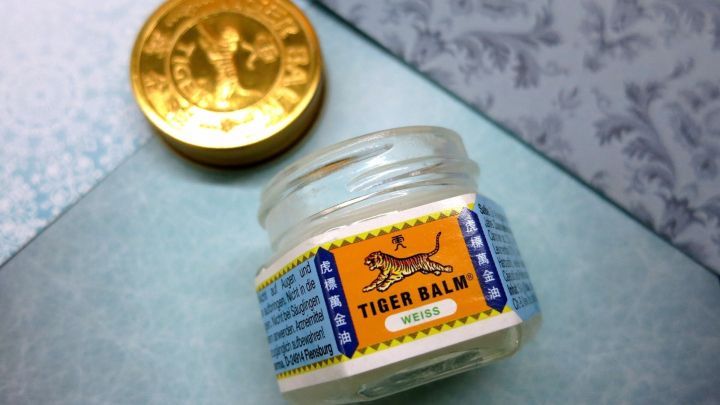 Ein leeres Glas Tiger Balm Weiss liegt offen neben dem goldfarbenen Deckel auf einer dekorativen Unterlage.