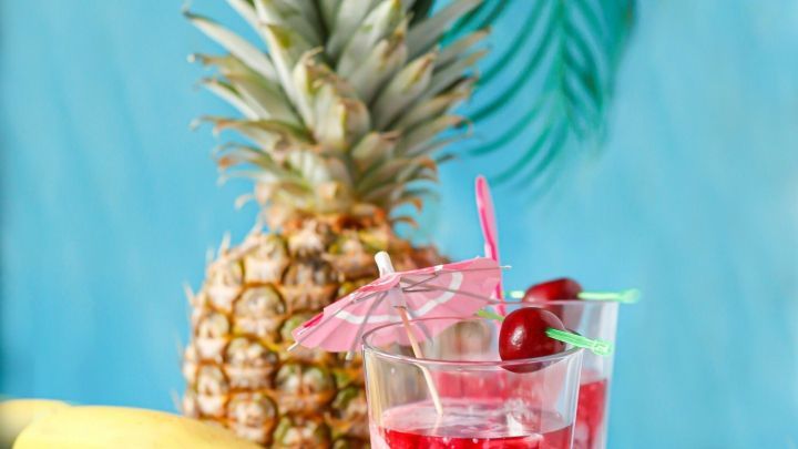 Ein frischer Banane-Kirsch Cocktail steht in Gl&auml;sern mit bunten Schirmchen, umgeben von Ananas und Bananen auf blauem Hintergrund.