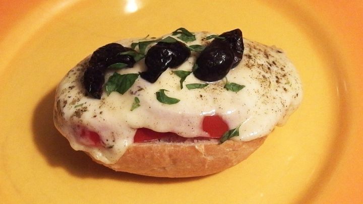 Ein Crostini mit geschmolzenem Mozzarella, Tomaten und Oliven auf einem gelben Teller, appetitlich garniert.