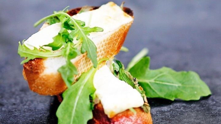 Ein ger&ouml;stetes Brotst&uuml;ck mit Rucola und Ziegenk&auml;se ist schmackhaft angerichtet auf einem dunklen, glatten Hintergrund.