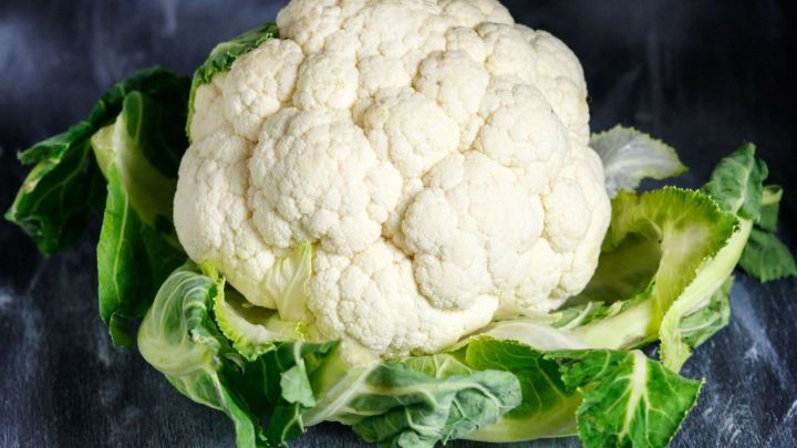 Ein frischer Blumenkohl liegt auf gr&uuml;nen Bl&auml;ttern und hebt sich vor einem dunklen Hintergrund hervor.