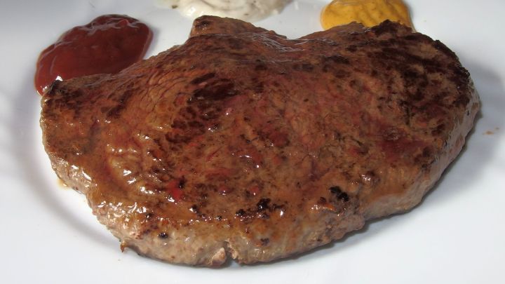 Ein z&auml;hes Steak liegt auf einem wei&szlig;en Teller, umgeben von Ketchup und Senf, bereit zum Verzehr.