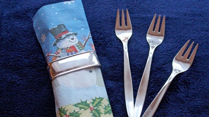 Eine Serviette mit einem Schneemannmotiv ist zusammengefaltet und wird von einer silbernen Serviettenring gehalten, neben drei silbernen Gabeln auf einem blauen Tisch.