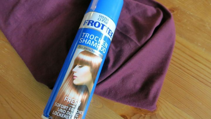 Eine Flasche Trockenshampoo liegt auf einem lila Kleidungsst&uuml;ck auf einem Holztisch, um Fettflecken zu entfernen.