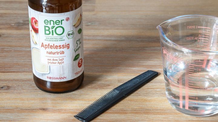 Eine Flasche mit Apfelessig und einem Kamm liegen neben einem Glas Wasser auf einem Holztisch, bereit zur Anwendung gegen Kopfl&auml;use.