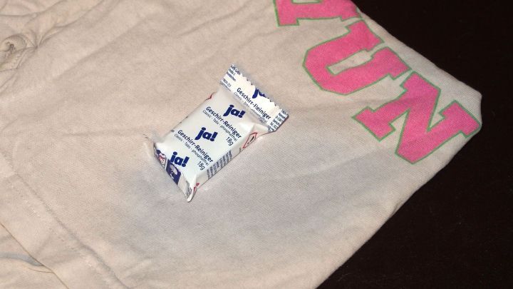 Ein beiges T-Shirt liegt auf einem dunklen Untergrund, daneben ein Sp&uuml;lmaschinentab, bereit zum Entfernen von Kaffeeflecken.