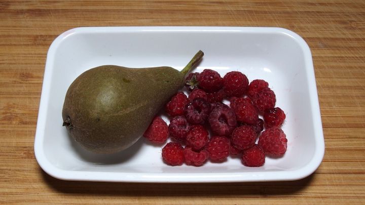 Eine gr&uuml;ne Birne liegt neben einer Gruppe roter Himbeeren auf einem wei&szlig;en Teller, auf einem Holzuntergrund.