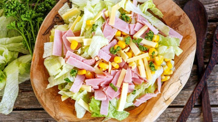 Frischer Chefsalat aus gr&uuml;nem Salat, Schinken, K&auml;se und Mais in einer Holzsch&uuml;ssel, umgeben von frischen Kr&auml;utern.