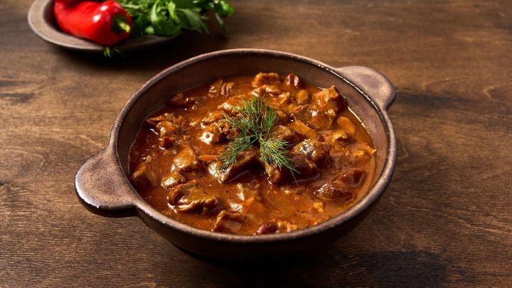Ein bauchiger Topf mit Sauerbratengulasch steht auf einem Holztisch, umgeben von frischem Koriander und Paprika.