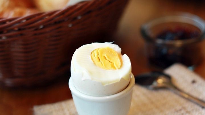 Ein perfekt gekochtes Fr&uuml;hst&uuml;cksei steht in einem Eierbecher, umgeben von frischem Brot und Marmelade auf einem Tisch.