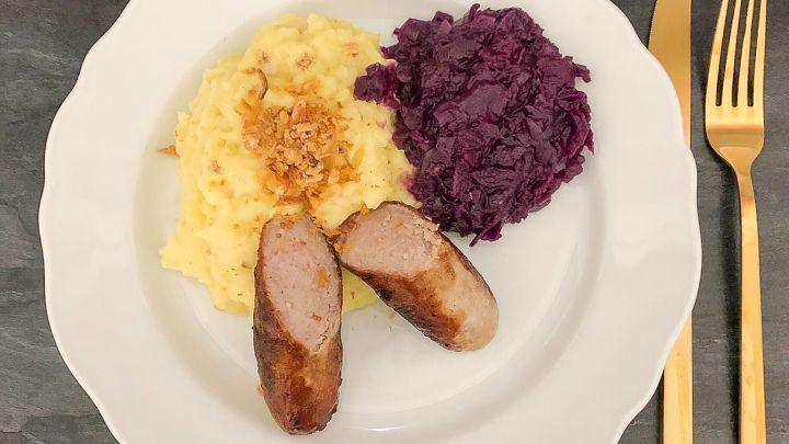 Das Kartoffelp&uuml;ree liegt saftig auf dem Teller, begleitet von Bratwurst und lila Rotkohl, umgeben von eleganten Bestecken.