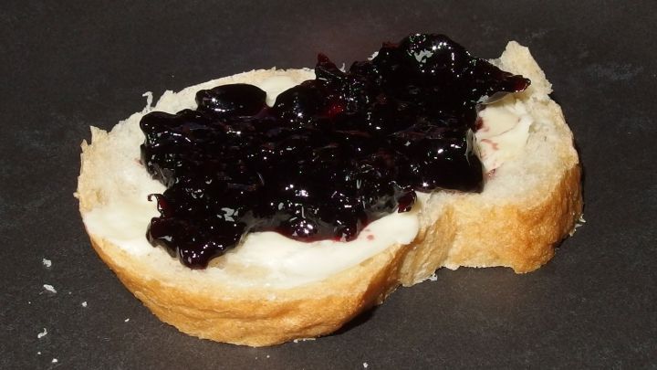 Ein St&uuml;ck Brot liegt auf einer dunklen Unterlage, belegt mit s&uuml;&szlig;er Johannisbeer-Kirschmarmelade.