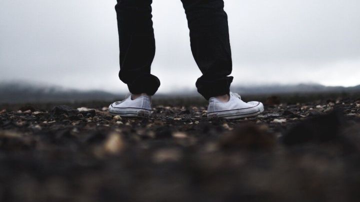 Wei&szlig;e Stoffturnschuhe stehen auf einem schmutzigen Untergrund, w&auml;hrend eine graue, wolkige Landschaft im Hintergrund sichtbar ist.