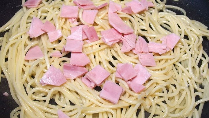 Butternudeln liegen in einer Pfanne, vermischt mit kleinen St&uuml;ckchen Schinken, umgeben von einem dunklen Hintergrund.