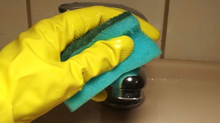 Eine Hand mit einem gelben Gummi-Handschuh reinigt eine gl&auml;nzende Armatur mit einem blauen Schwamm in einer sp&uuml;len&auml;hnlichen Umgebung.