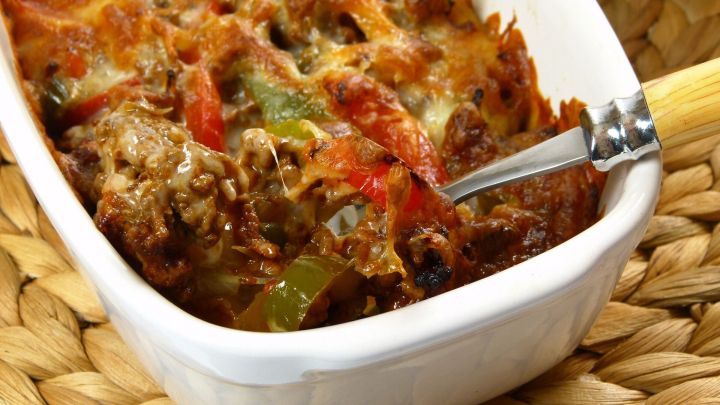 Eine gebackene Mischungsplatte mit gebratenem Fleisch, Gem&uuml;se und K&auml;se wird mit einem L&ouml;ffel aus einer wei&szlig;en Auflaufform serviert.