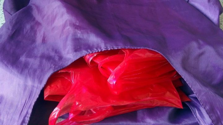 Eine lila Einkaufstasche mit roten Plastikt&uuml;ten, die darin verstaut sind, steht auf einer hellen Oberfl&auml;che.