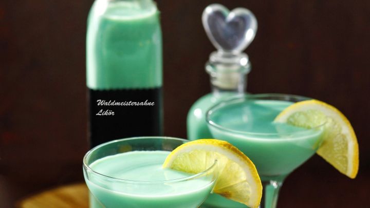 Zwei Glasbeh&auml;lter mit mintgr&uuml;nem Waldmeistersahnelik&ouml;r stehen auf einem Holzuntergrund, dekoriert mit Limettenscheiben.