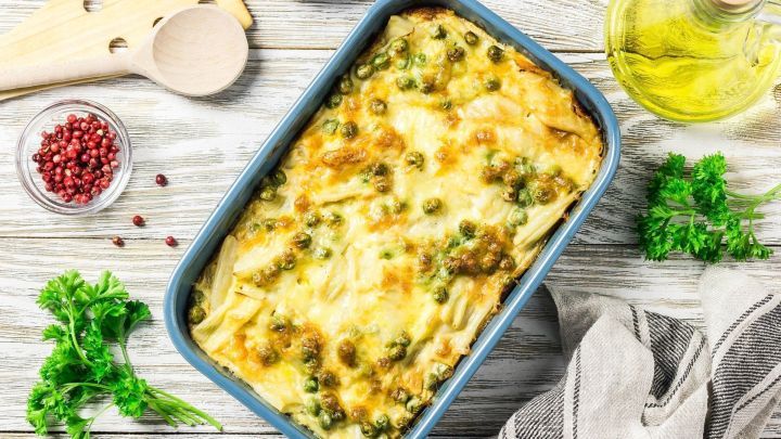 Ein appetitlicher Nudelauflauf mit K&auml;se und Erbsen wird in einer blauen Auflaufform auf einem Holztisch pr&auml;sentiert, umgeben von Zutaten.