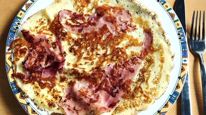 Eine gro&szlig;e Pfannkuchen-Pizza liegt auf einem bunten Teller, belegt mit knusprigem Speck, auf einem Holztisch.