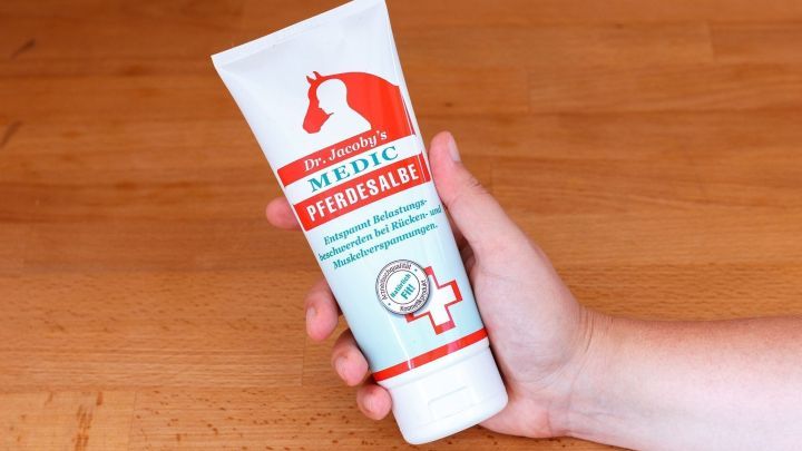 Eine Hand h&auml;lt eine Tube Pferdebalsam, w&auml;hrend sie auf einem h&ouml;lzernen Tisch steht, um gegen Prellungen zu helfen.