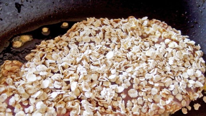 Ein panierter Schnitzel liegt in einer Pfanne, umgeben von goldbraun ger&ouml;steten Haferflocken in hei&szlig;em &Ouml;l.