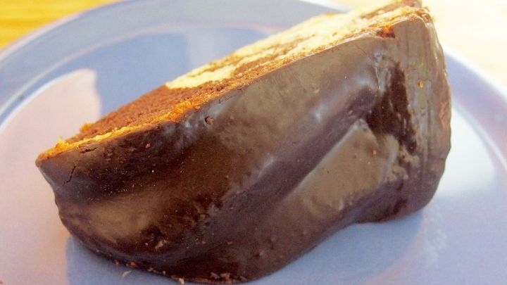 Ein St&uuml;ck Kuchen, &uuml;berzogen mit geschmolzener Schokoladenglasur, liegt auf einem hellblauen Teller in einer K&uuml;che.