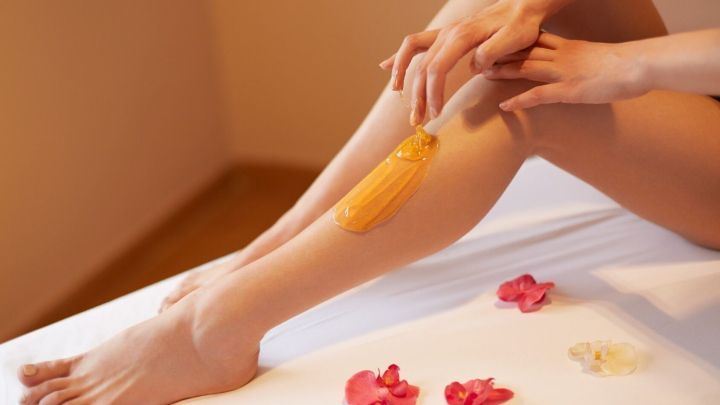 Eine Person entfernt mit einer selbstgemachten Zuckerpaste Haare von ihrem Bein, umgeben von Bl&uuml;tenbl&auml;ttern auf einem hellen Untergrund.