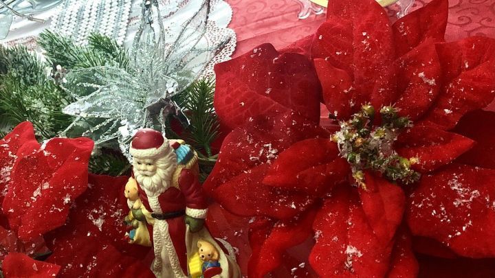 Ein festlicher Dekoschneee ist um eine rote Weihnachtssternpflanze arrangiert, mit Santa-Figur und Glitzer im Hintergrund.