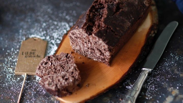 Ein dunkles Schokobrot liegt auf einem Holzbrett, mit einem St&uuml;ck abgeschnitten, umgeben von Mehlresten.