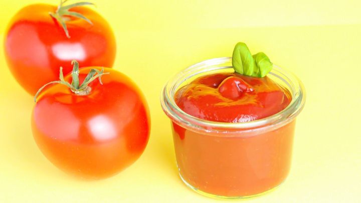 Rote Tomaten stehen neben einem Glas Ketchup, das mit frischem Basilikum dekoriert ist, auf gelbem Hintergrund.