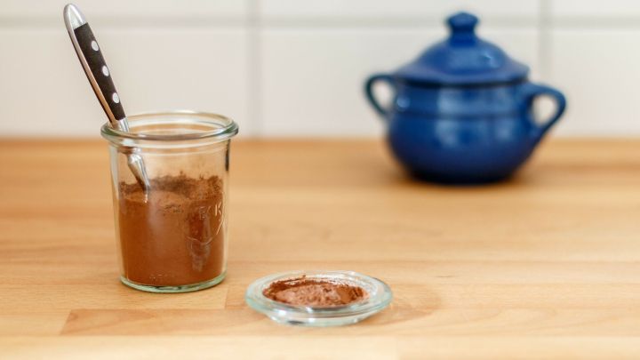 Ein Glas mit Backkakao steht auf einem Holztisch, w&auml;hrend ein blauer Topf im Hintergrund sichtbar ist.