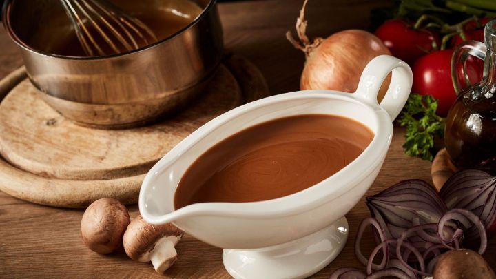 Eine wei&szlig;e So&szlig;ensch&uuml;ssel mit saurer So&szlig;e steht auf einem Holztisch, umgeben von frischem Gem&uuml;se und einer R&uuml;hrsch&uuml;ssel.