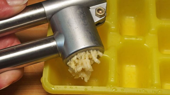 Eine Knoblauchpresse dr&uuml;ckt frischen Knoblauch in eine gelbe Eisw&uuml;rfelschale zur sp&auml;teren Verwendung.