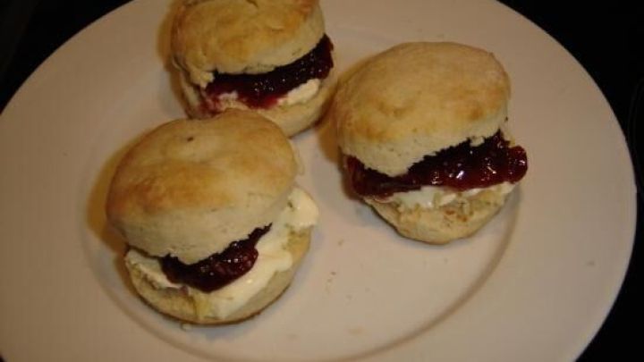 Drei englische Scones gef&uuml;llt mit dunkler Marmelade und Sahne liegen auf einem wei&szlig;en Teller, der in einem dunklen Raum steht.