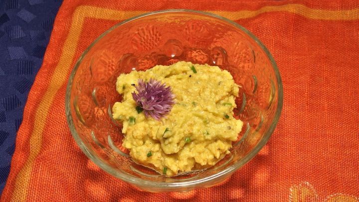 Ein Glas mit cremigem Avocado-Brotaufstrich steht auf einem orangefarbenen Tuch und ist mit einer essbaren Blume dekoriert.