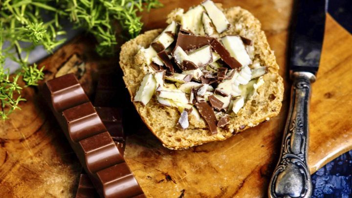 Ein belegtes Br&ouml;tchen mit gehackter Schokolade liegt auf einem Holzbrett, umgeben von frischen Kr&auml;utern.