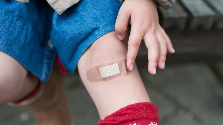 Ein Kind ber&uuml;hrt sanft ein Pflaster auf dem Knie, w&auml;hrend es auf einer Holzbank sitzt, umgeben von nat&uuml;rlicher Umgebung.