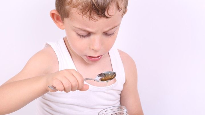 Kind l&ouml;ffelt honigartige Fl&uuml;ssigkeit, die aus Glas tropft, Auge auf L&ouml;ffel gerichtet, Hintergrund wei&szlig;.