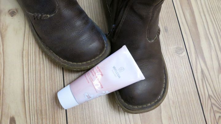 Braune Stiefel stehen neben einer Tube Reinigungsmilch auf einem Holzfu&szlig;boden, bereit zum Putzen und Pflegen.