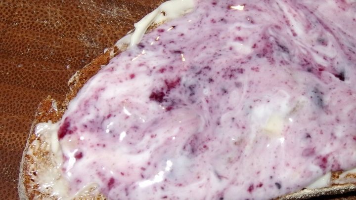 Ein Brotst&uuml;ck ist mit fruchtigem Joghurt bestrichen, auf einer rustikalen Holzunterlage, bereit zum Fr&uuml;hst&uuml;ck.