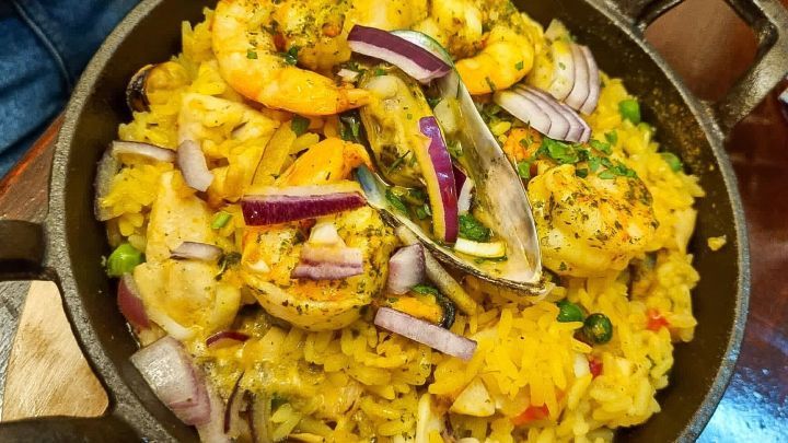 Eine Pfanne zeigt aromatische Paella mit Meeresfr&uuml;chten, gekr&ouml;nt von Garnelen und Gem&uuml;se, in einem rustikalen Restaurant.